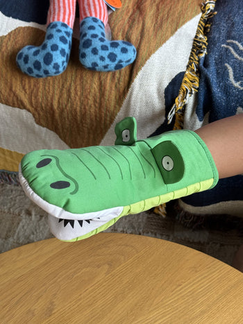 A person wears the Fluid Market Crocodile Oven Mitt by Fluid Market on their hand above a light wooden surface, with a blue spotted stuffed animal and a brown patterned blanket in the background.