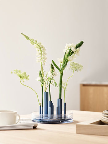 A Fritz Hansen Ikeru Low Vase with a floral arrangement of white flowers in its blue, multi-tubed circular structure on a wooden table.