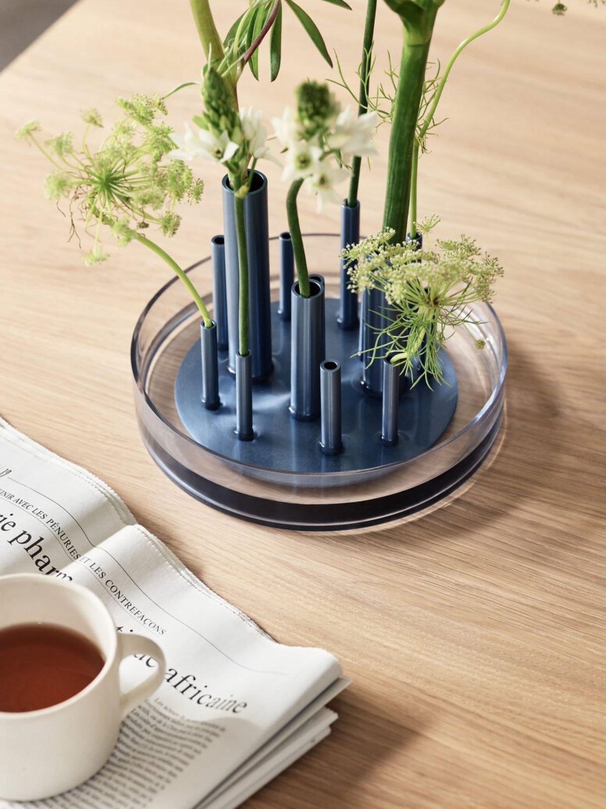 A circular blue Fritz Hansen Ikeru Low Vase holds multiple stems of green and white flowers, sitting on a wooden table next to a teacup and a newspaper.