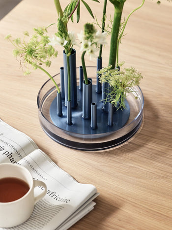 A circular blue Fritz Hansen Ikeru Low Vase holds multiple stems of green and white flowers, sitting on a wooden table next to a teacup and a newspaper.