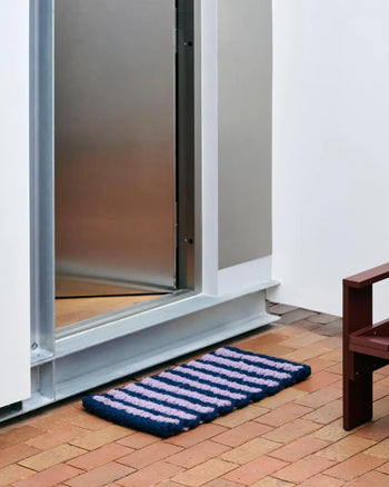 The HAY Coco Door Mat Stripe Wide by HAY, featuring blue and pink stripes and made from sturdy coir fibers, lies on a brick floor before a slightly open metal door with part of a brown bench visible on the right.