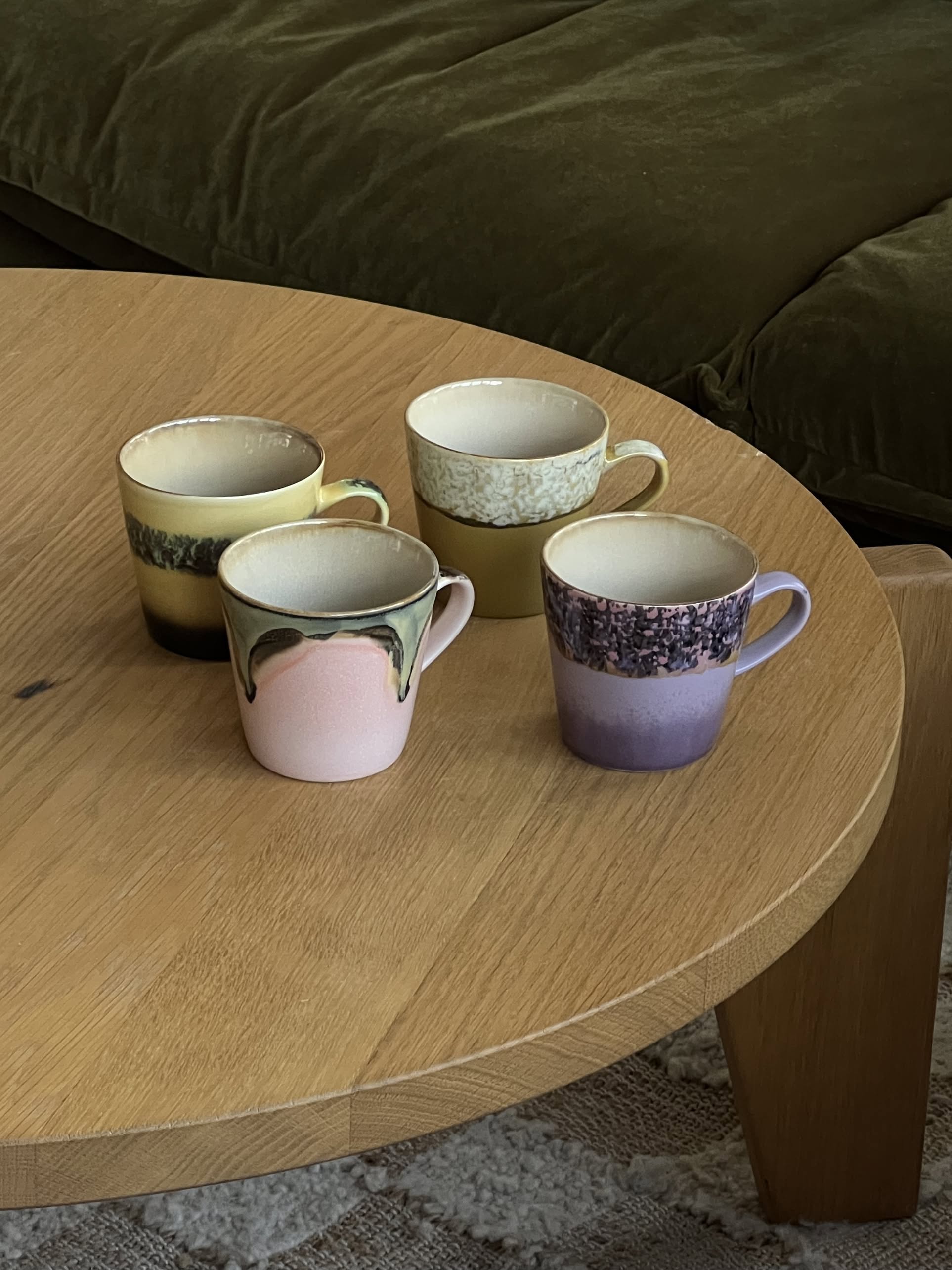 A set of four HKliving 70s Ceramics Americano Mugs, each with colorful glaze patterns, is arranged on a light wooden round table with a green couch partially visible in the background.