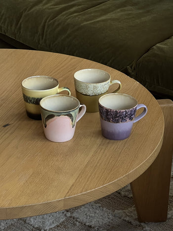 A set of four HKliving 70s Ceramics Americano Mugs, each with colorful glaze patterns, is arranged on a light wooden round table with a green couch partially visible in the background.