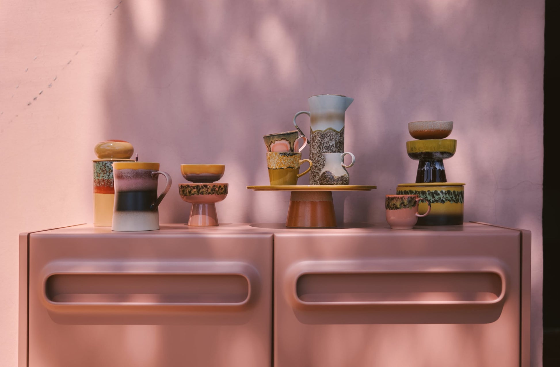 A pastel pink cabinet displays HKliving’s 70s Ceramics: Americano Mugs, Set of 4, with bowls, plates, and a pitcher. The scene is softly lit with dappled shadows and a pink wall background, highlighting the vintage mug design.