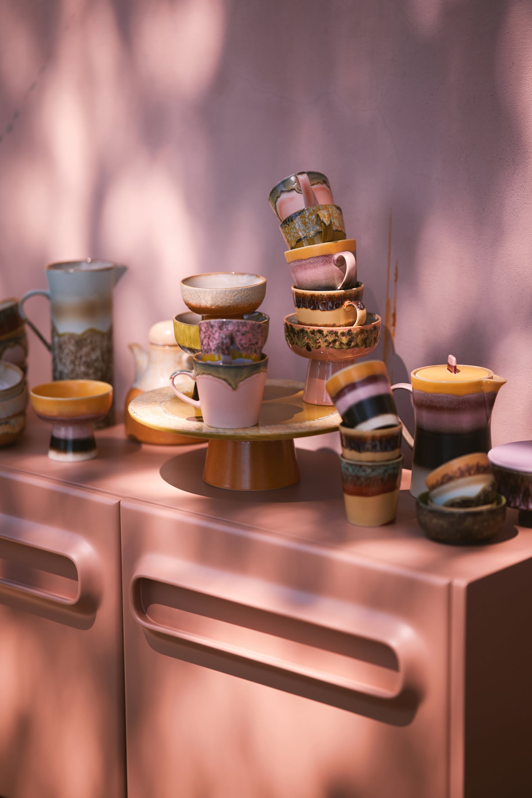 HKliving 70s Ceramics Cappuccino Mugs, Set of 4, are stacked on a pastel pink cabinet against a pink wall, with dappled sunlight and shadows lending a soft, artistic touch.