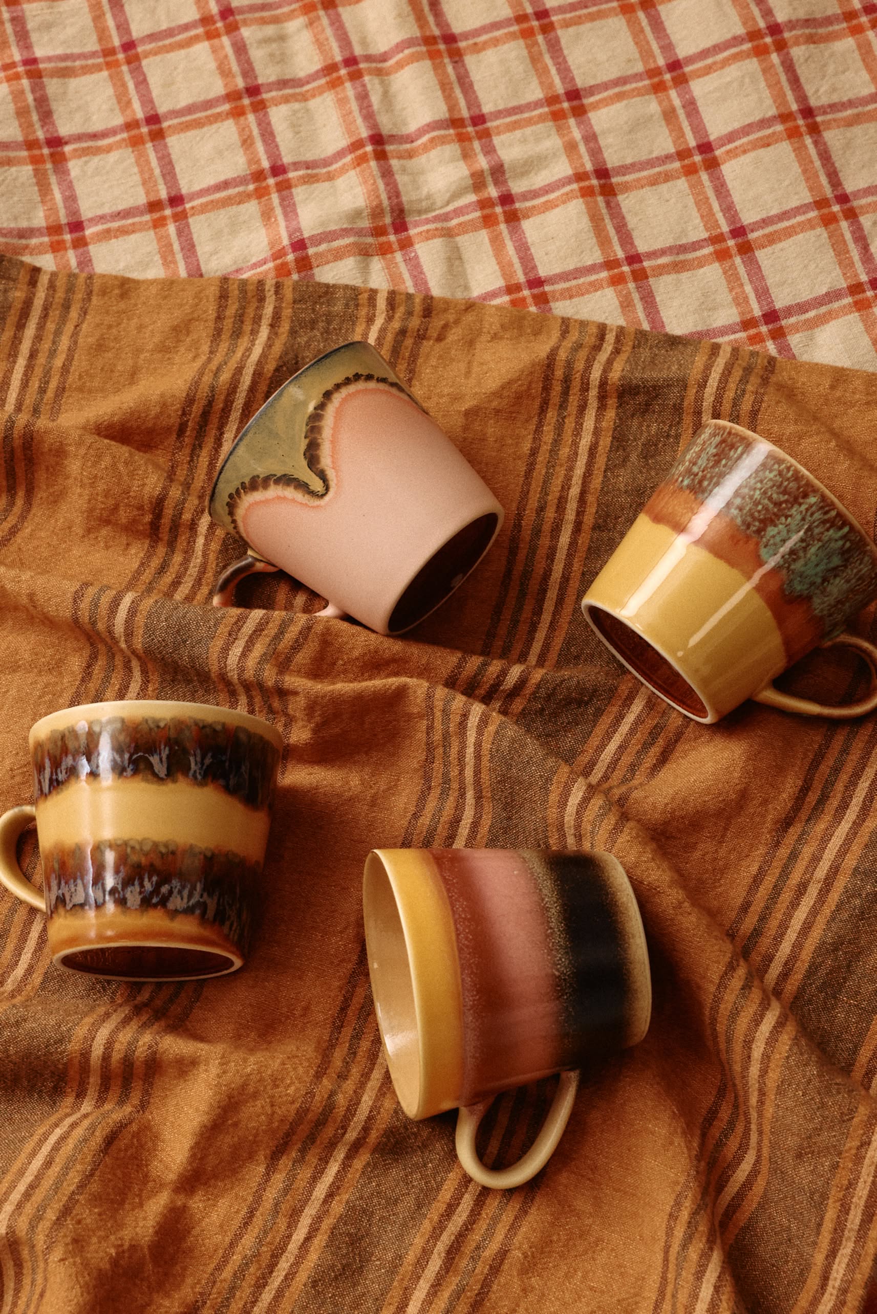 The HKliving 70s Ceramics Cappuccino Mugs, Set of 4, are shown in earthy tones with rustic patterns on brown and orange striped fabric, with a plaid cloth in the background.
