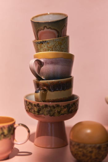 A stack of five colorful, mismatched retro mugs and bowls is balanced against a soft pink background, accompanied by an HKliving 70s Ceramics: Cappuccino Mugs (Set of 4) from HKliving and a round container nearby.