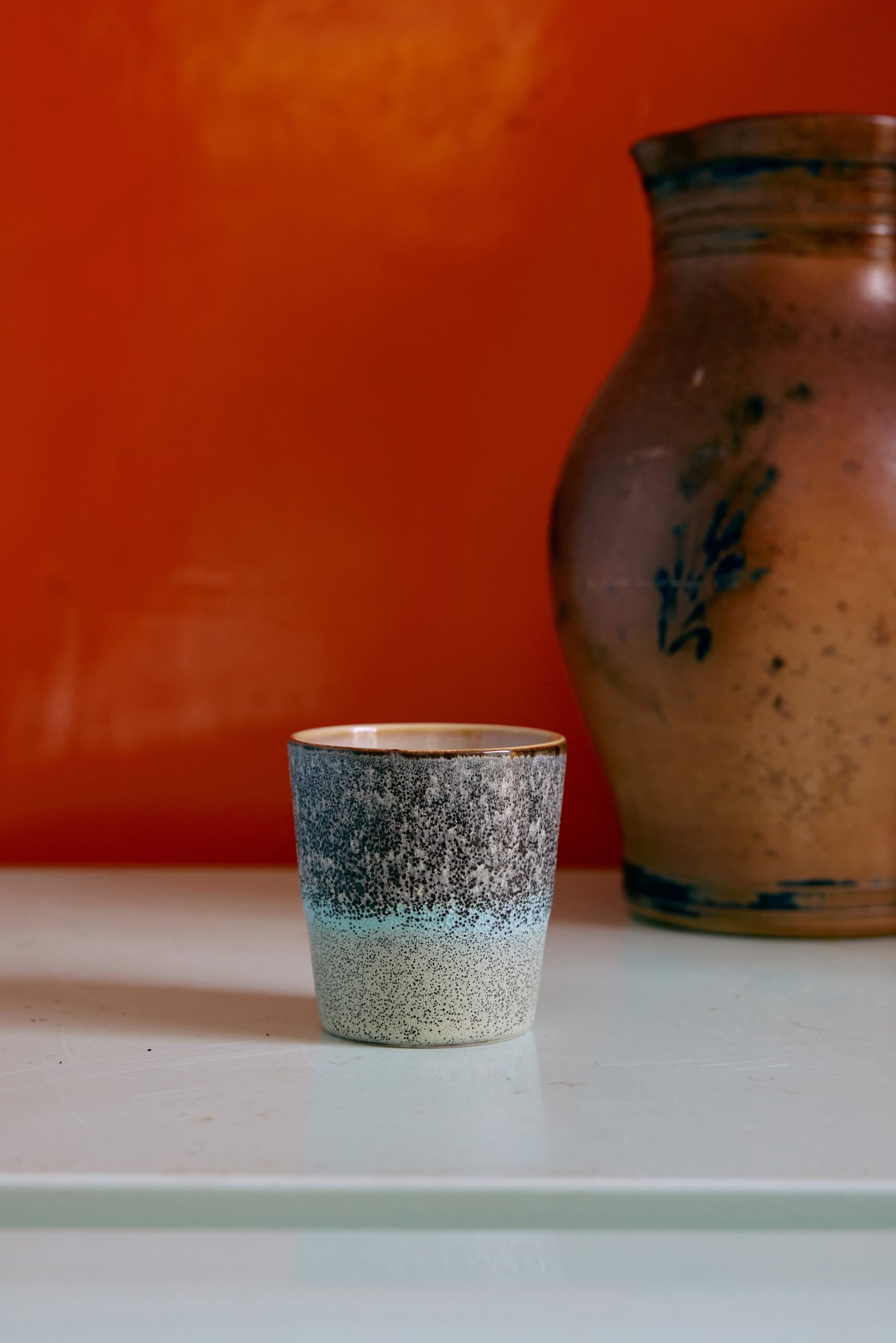 An HKliving 70s Ceramics coffee mug from a set of 6 sits on a light surface before an orange wall, next to a large brown clay vase with dark blue decoration.