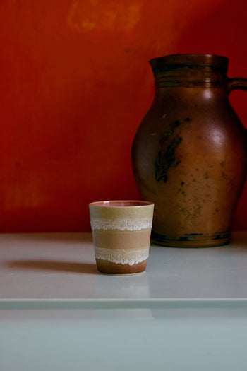 An HKliving 70s Ceramics Coffee Mug from the set of 6 rests on a light surface in front of a large, rustic brown jug, with a bold reddish-orange wall in the background.