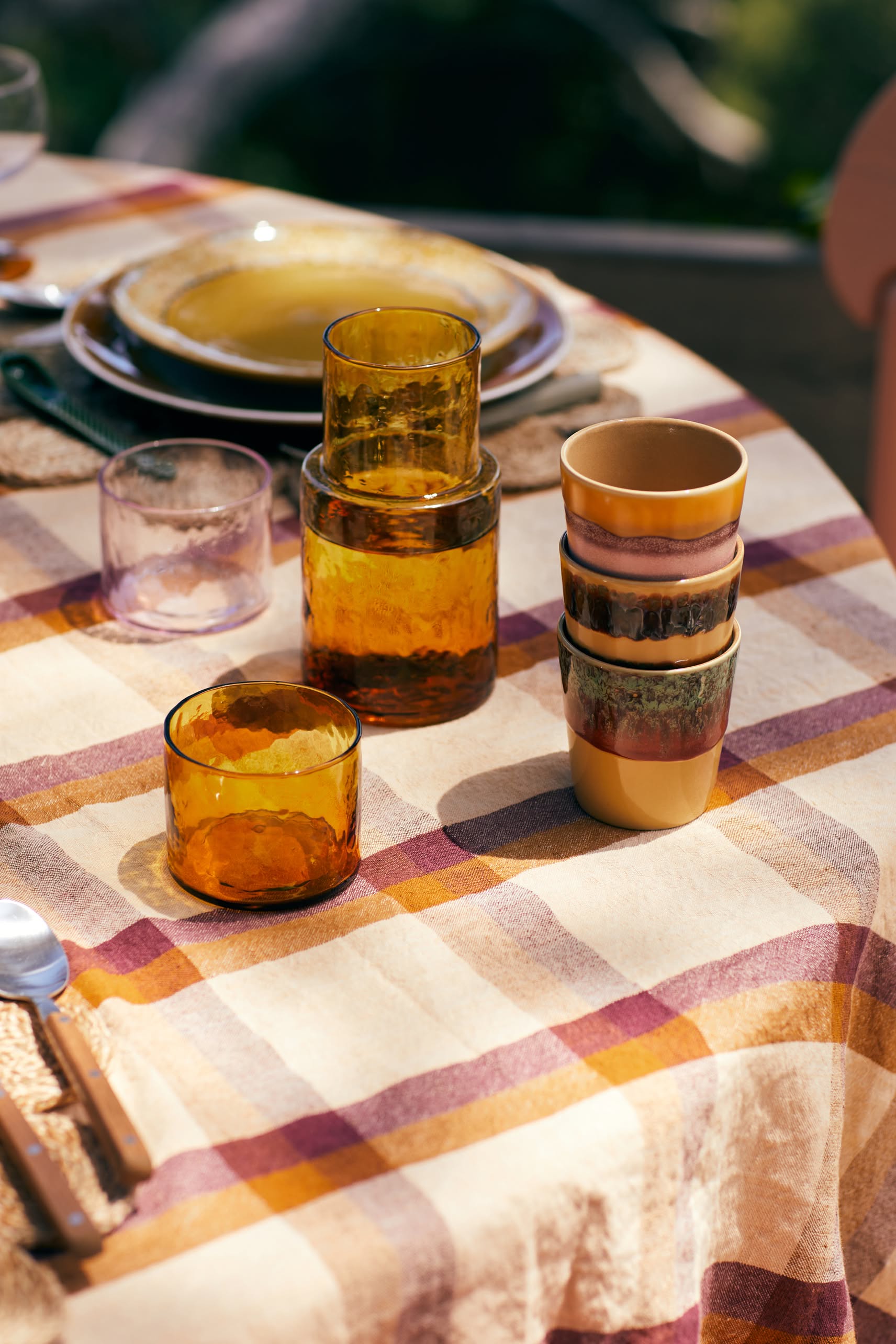 A sunlit table is set with a plaid tablecloth, stacked amber glasses, cutlery, plates, and HKliving 70s Ceramics Coffee Mugs (Set of 6) by HKliving, creating a warm and inviting outdoor dining scene.