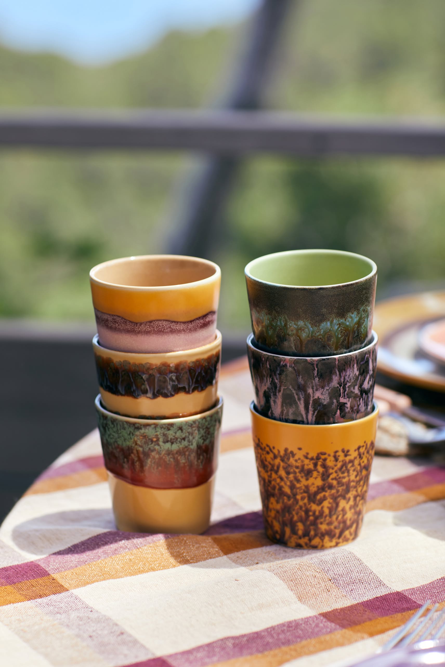 The HKliving 70s Ceramics: Coffee Mugs, Set of 6, features vibrant, uniquely patterned mugs stacked outdoors on a checkered tablecloth, capturing vintage 70s style with a blurred green background.
