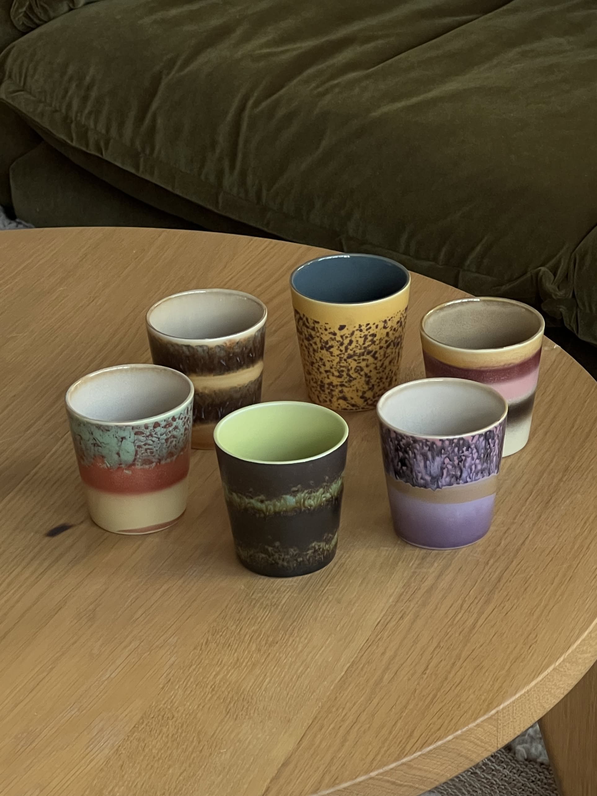 The HKliving 70s Ceramics: Coffee Mugs, Set of 6, by HKliving features colorful mugs with unique patterns and textures arranged on a light wooden table, with a dark green sofa in the background.