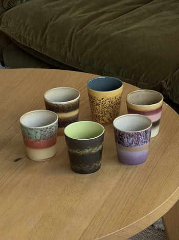 The HKliving 70s Ceramics: Coffee Mugs, Set of 6, by HKliving features colorful mugs with unique patterns and textures arranged on a light wooden table, with a dark green sofa in the background.
