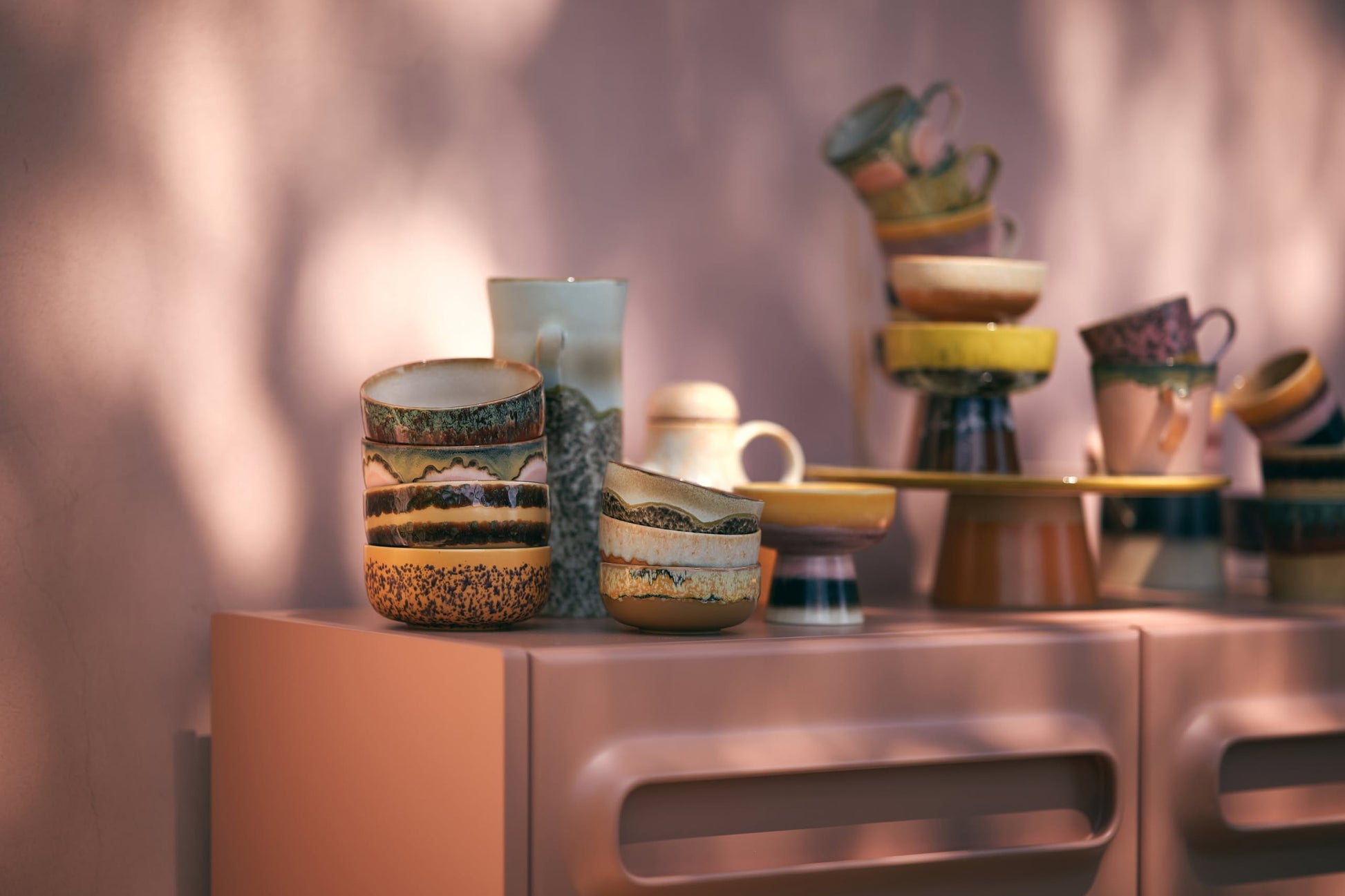 HKliving 70s Ceramics: Dessert Bowls, Set of 4, featuring vibrant patterns, are displayed on a light pink cabinet. Gentle sunlight adds soft shadows and warmth to the cozy setting.