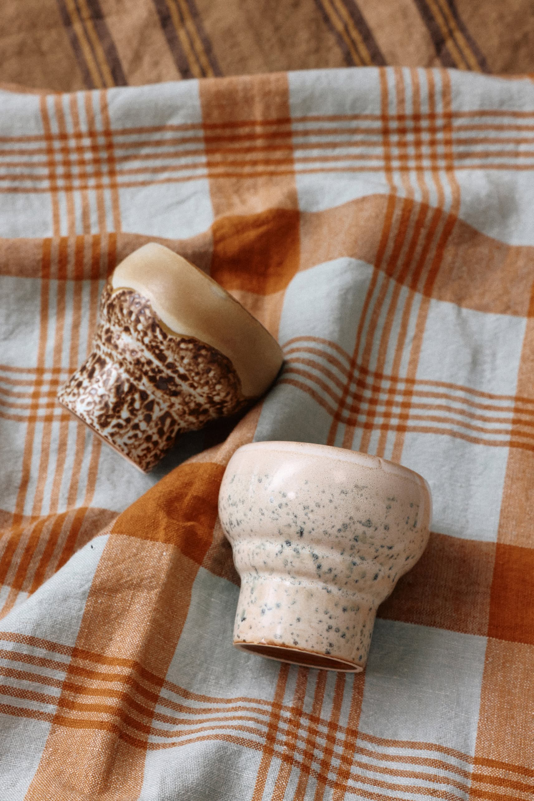 The HKLIVING 70s Ceramics: Lungo Mugs, Set of 2 by HKLIVING are small, textured mugs ideal for coffee lovers, shown upside down on a blue, beige, and orange plaid fabric.
