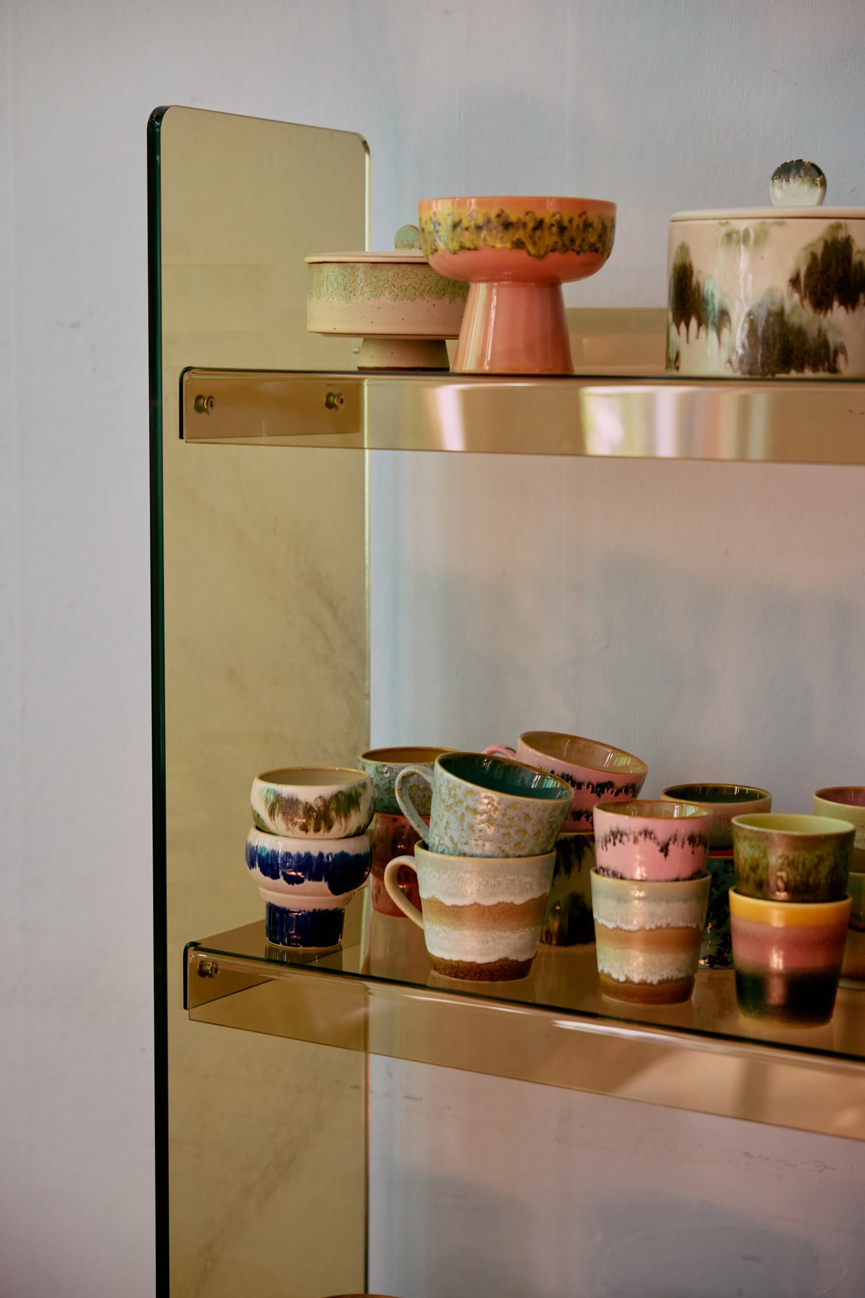 A glass and gold shelf showcases HKliving 70s Ceramics Lungo Mugs—set of 2—with unique textures, retro patterns, and earthy tones like green, blue, pink, and brown.