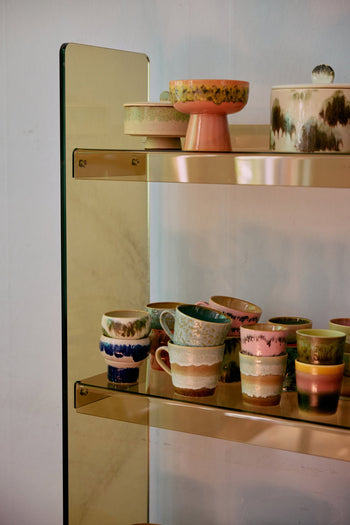 A glass and gold shelf showcases HKliving 70s Ceramics Lungo Mugs—set of 2—with unique textures, retro patterns, and earthy tones like green, blue, pink, and brown.