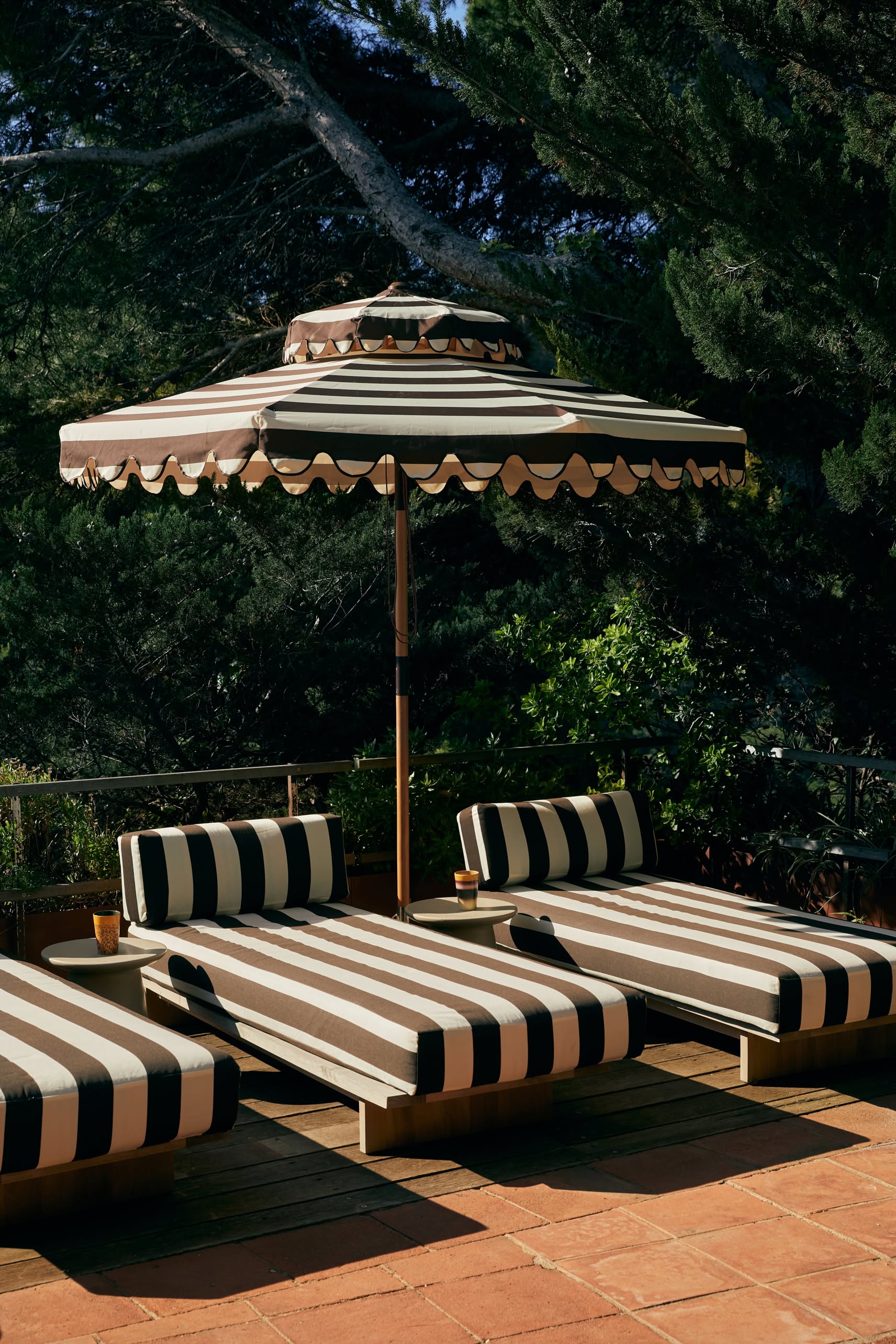 Three black and white striped lounge chairs with matching cushions sit under a large striped umbrella on a terracotta patio, with an HKliving 70s Ceramics: Tea Mugs Set of 2 by HKliving resting nearby, surrounded by lush greenery.