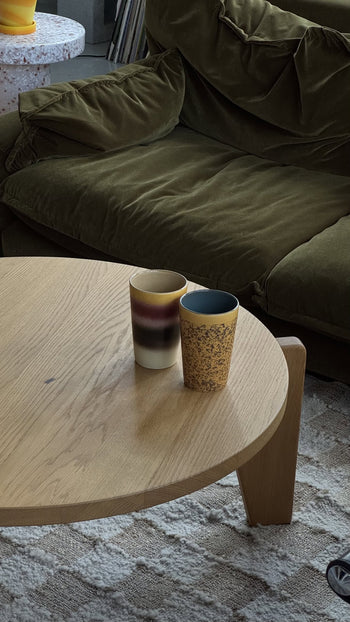Two HKliving 70s Ceramics tea mugs, set of 2, rest on a round wooden coffee table before a green velvet sofa. Each mug features a unique color, with soft lighting creating a cozy atmosphere.