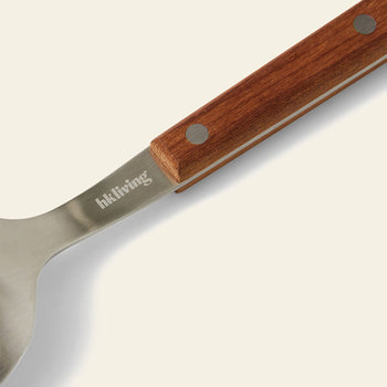 Close-up of a utensil from the HKLIVING 70s Cutlery: Wood, Set of 16, featuring a stainless steel head, wooden handle with two metal rivets, and HKLIVING engraved on the metal.