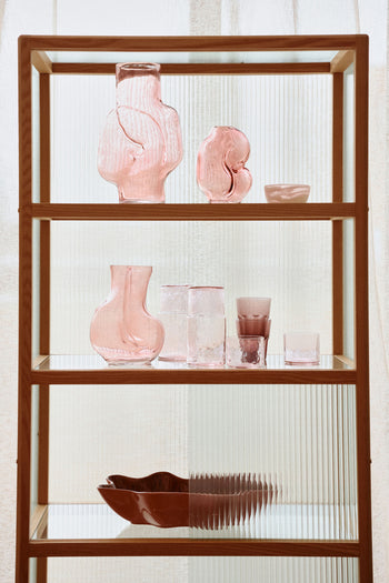 A wooden and glass shelf displays HKLIVING Gleam Glass Vases in pink, clear, and olive green, each with unique artistic shapes. Sunlight filters through sheer curtains behind them.