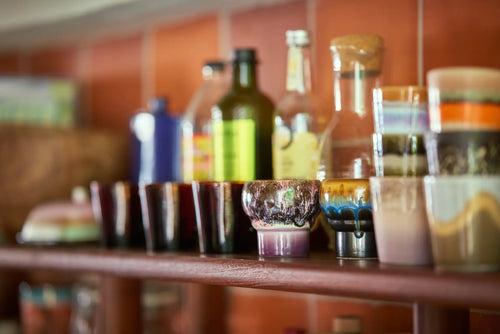 A close-up of HKLIVING 70s Ceramics: Lungo Mugs (Set of 2) and colorful glass cups on a wooden shelf, with more bottles and objects blurred in the background against a brown tiled wall.