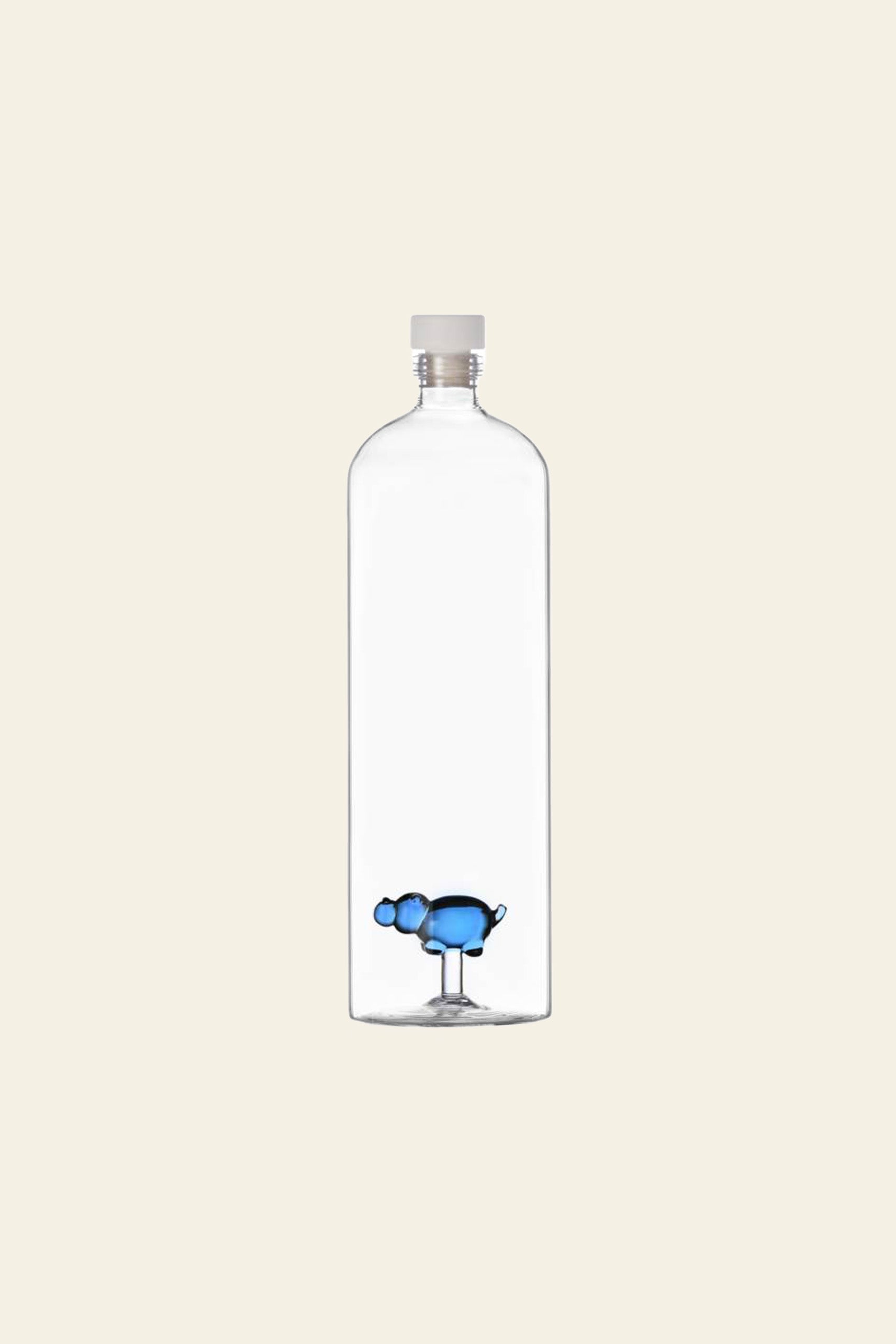 A clear, hand-crafted glass bottle with a white cap features a small blue hippo figure at the base—a whimsical design from Ichendorf Milano’s Animal Farm Bottle Big collection. The background is plain and off-white.