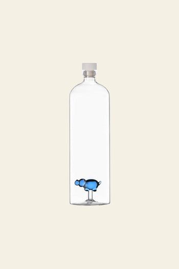 A clear, hand-crafted glass bottle with a white cap features a small blue hippo figure at the base—a whimsical design from Ichendorf Milano’s Animal Farm Bottle Big collection. The background is plain and off-white.