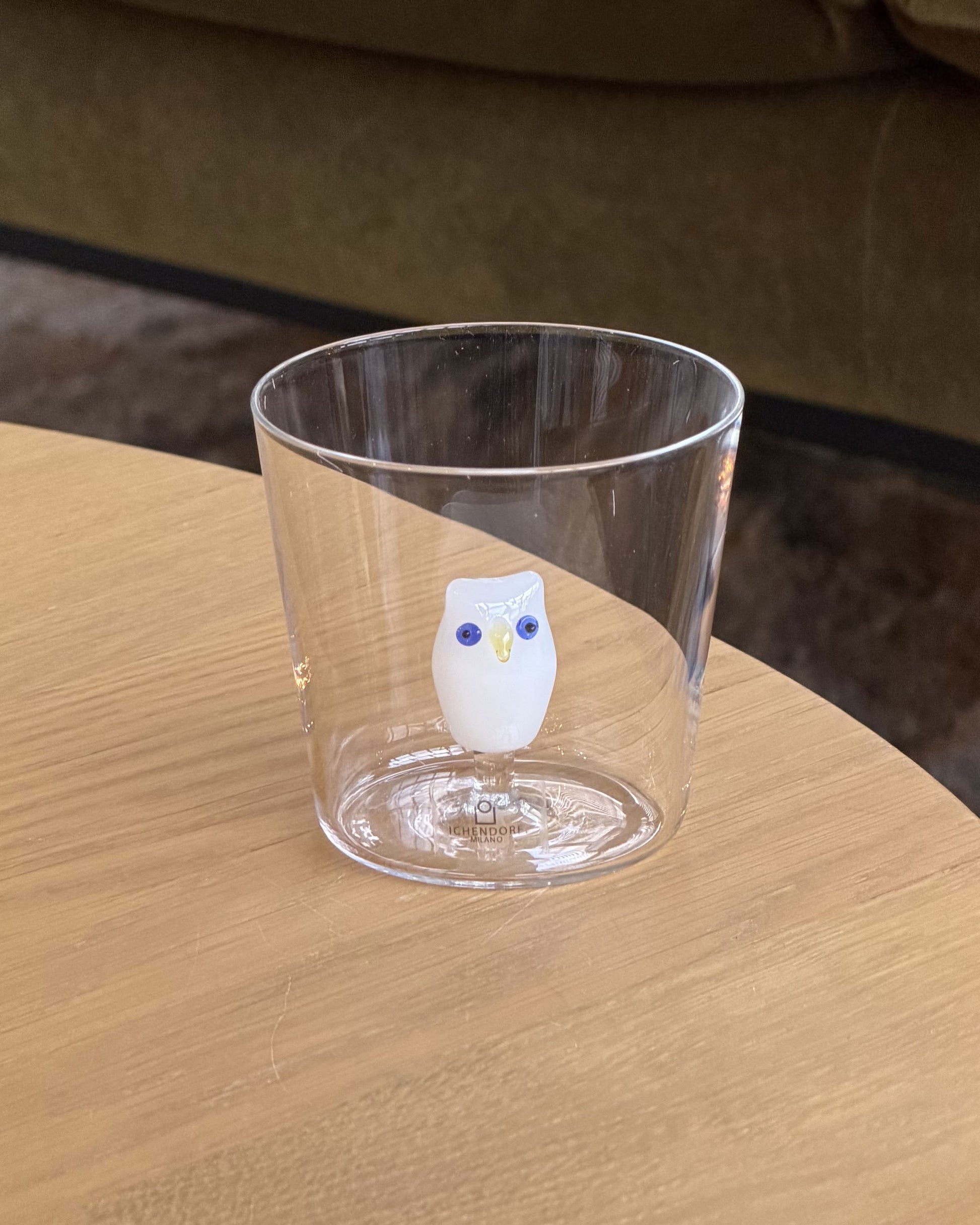 The Ichendorf Milano Animal Farm Tumbler, a clear glass by Ichendorf Milano, features a small white owl figurine and is shown on a light wooden surface with a green couch in the background.