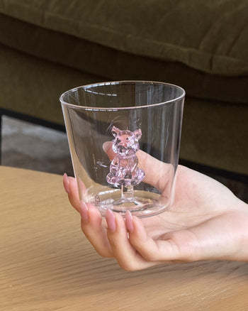 A manicured hand holds an Ichendorf Milano Animal Farm Tumbler, which features a small pink glass bear at its base. A wooden surface and green sofa are visible in the background.