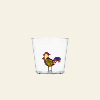 The Ichendorf Milano Animal Farm Tumbler by Ichendorf Milano is a clear borosilicate glass tumbler featuring a colorful rooster figurine with red, yellow, and blue accents at the base, set against a plain off-white background.