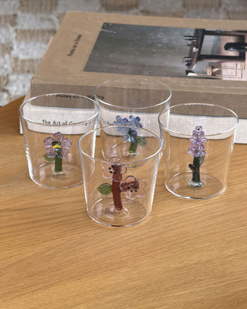 Four clear Ichendorf Milano Botanica Tumblers, each handmade with a unique colorful floral design—including the Light Blue Flower—are displayed on a wooden table. A beige-covered book rests in the background.