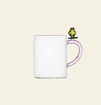 The Ichendorf Milano Fruits and Flower Mug is a clear glass mug with a light pink handle, adorned by a small green and yellow frog figurine designed by Alessandra Baldereschi for Ichendorf Milano.