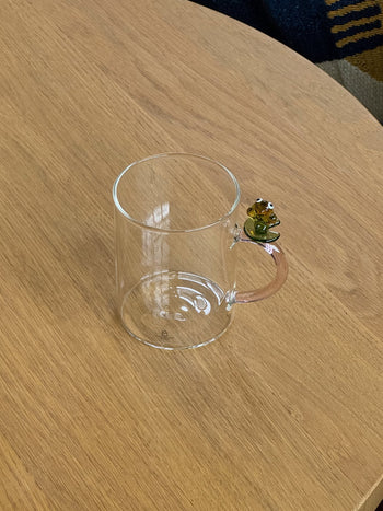 The Ichendorf Milano Fruits and Flower Mug by Ichendorf Milano, with a decorative frog on the handle, rests on a light wooden table.