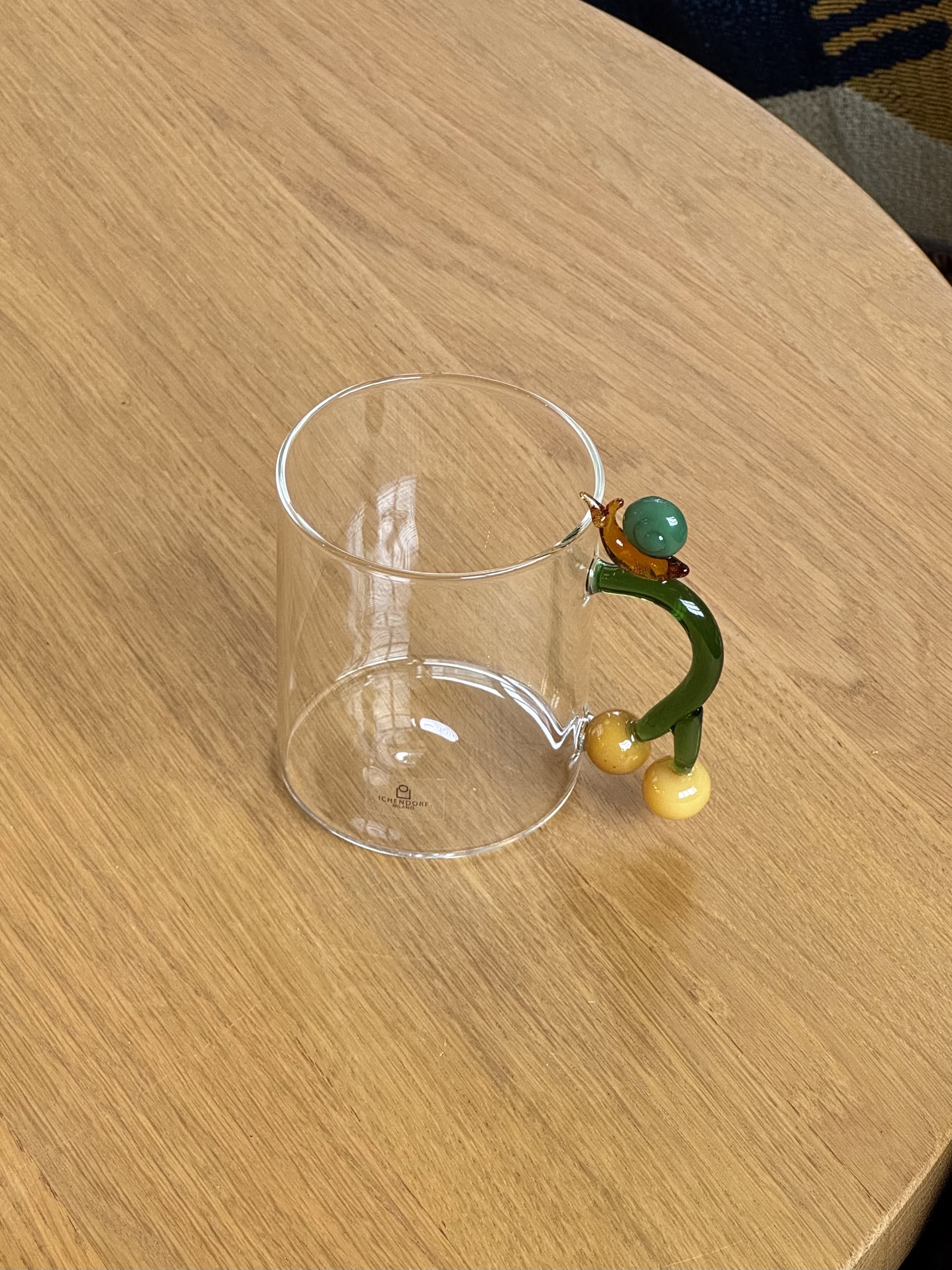 The Ichendorf Milano Fruits and Flower Mug, crafted in clear glass with a whimsical green handle, sits on a light wooden table.