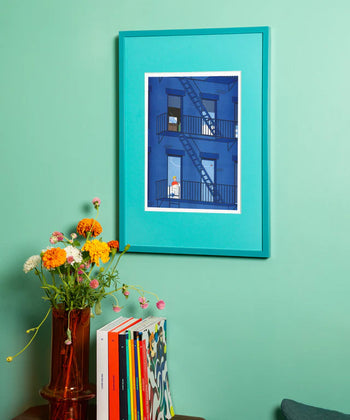 The KIBLIND Atelier Simon Bailly - Porco Rosso poster by KIBLIND Atelier, inspired by Franco-Belgian comics, shows a blue building with fire escapes and hangs above a small table with flowers and colorful books.