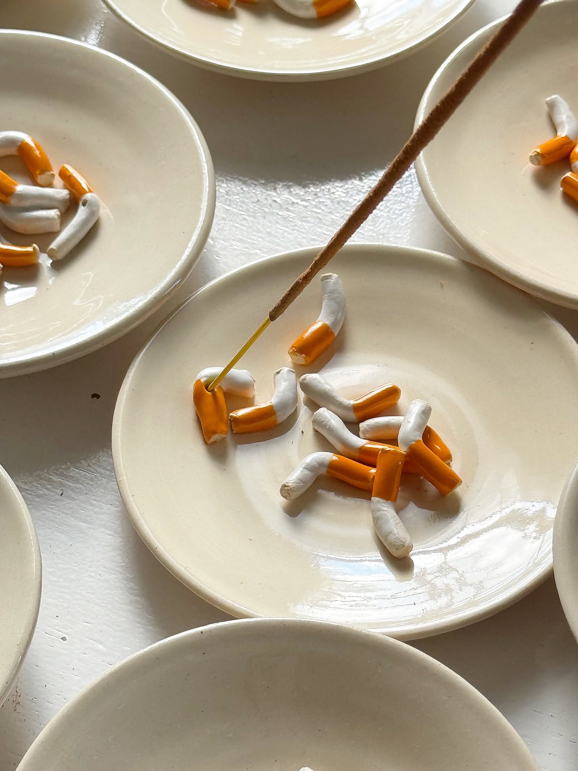 The MIRYO Cigarette Incense Holder by MIRYO features a beige plate with clay cigarette butts that hold incense sticks, blending their ash with the sculptural design. Similar holders can be seen alongside it for a unique display.