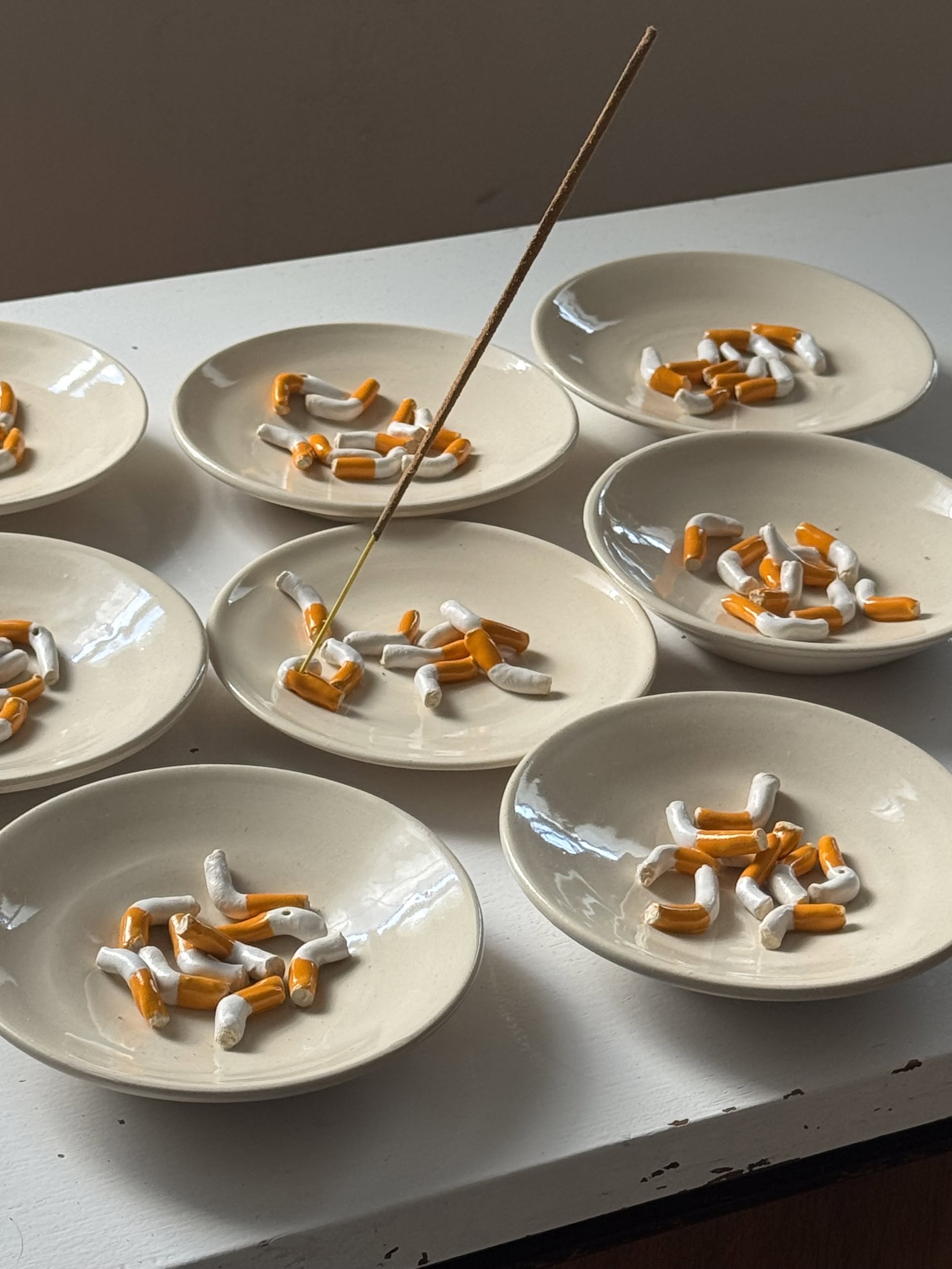 Nine small white plates, each with broken cigarette pieces, are arranged on a white surface. One plate features the MIRYO Cigarette Incense Holder by MIRYO, holding a lit incense stick among the cigarette pieces under soft light.