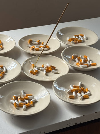 Nine small white plates, each with broken cigarette pieces, are arranged on a white surface. One plate features the MIRYO Cigarette Incense Holder by MIRYO, holding a lit incense stick among the cigarette pieces under soft light.