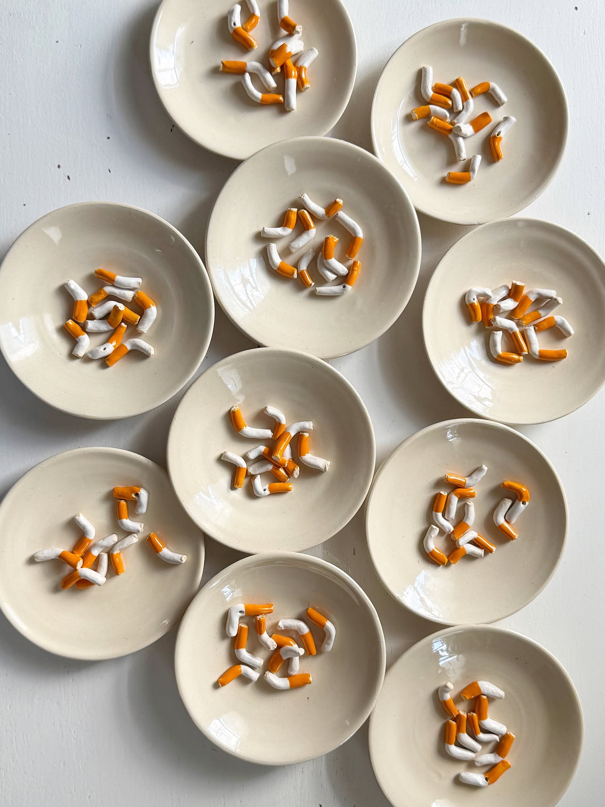 Eleven beige MIRYO Cigarette Incense Holders by MIRYO are arranged on a white surface, each with orange and white elements resembling cigarette butts—a sculptural and unique incense holder display.