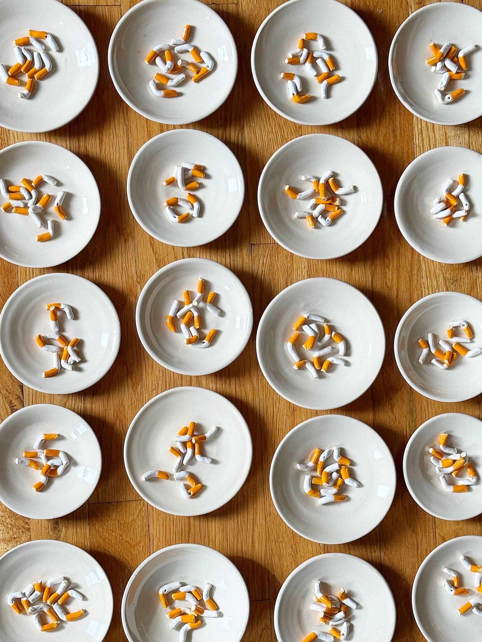 Twenty MIRYO Cigarette Incense Holders by MIRYO are arranged in a grid on a wooden surface, each resembling broken cigarette butts with white filters and orange tips, creating a conceptual art display.