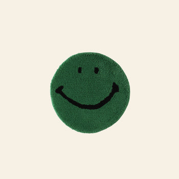 The MIYOSHI RUG Harvey Ball x MIYOSHI RUG Original Smiley Face Chair Rug features a green circular design with a simple black smiley face, inspired by Harvey Ball, on a light beige background.