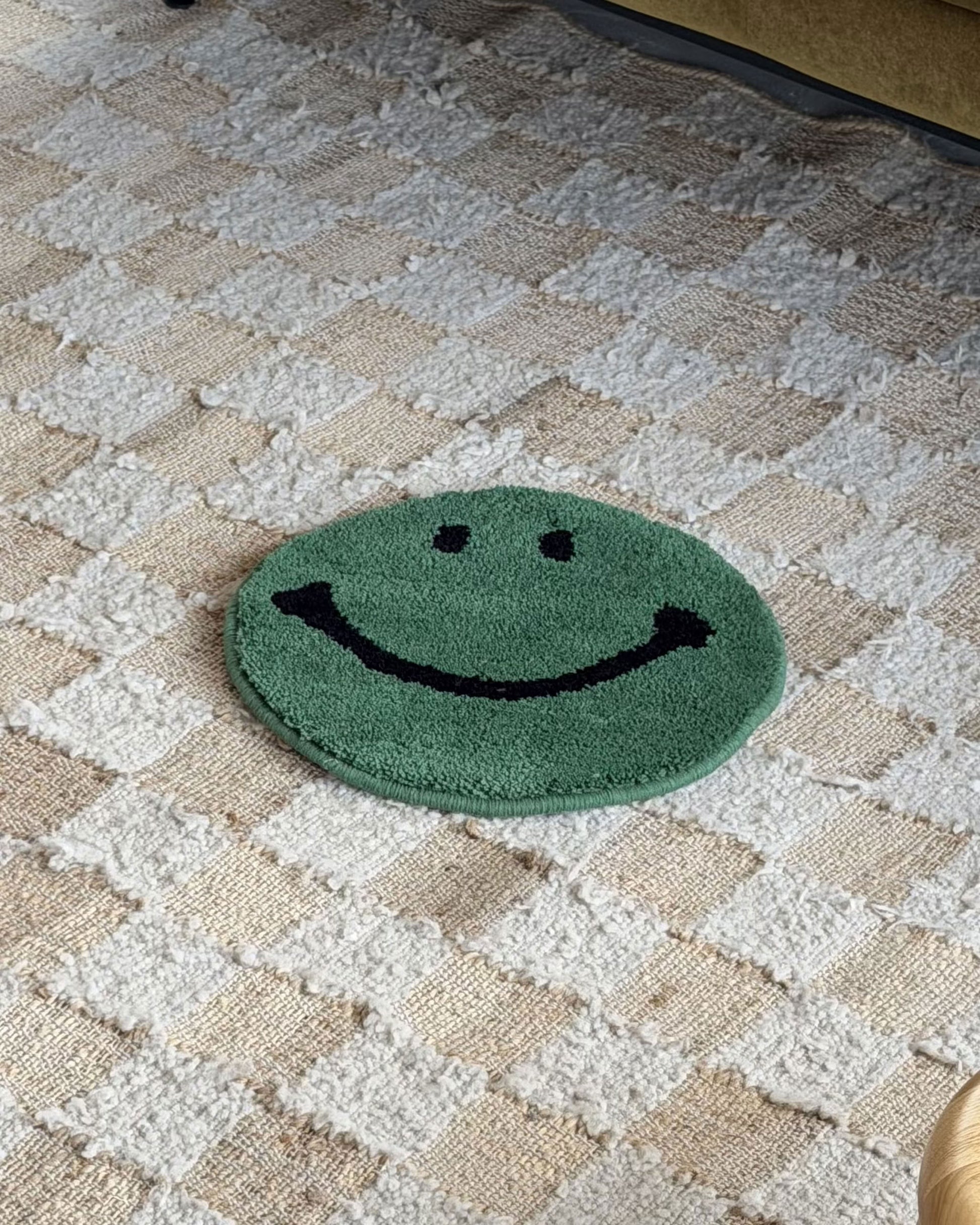 The MIYOSHI RUG Harvey Ball x MIYOSHI RUG Original Smiley Face Chair Rug, a small round green rug inspired by Harvey Ball and handmade in Japan, rests on a textured beige and white checkered carpet.