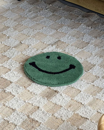 The MIYOSHI RUG Harvey Ball x MIYOSHI RUG Original Smiley Face Chair Rug, a small round green rug inspired by Harvey Ball and handmade in Japan, rests on a textured beige and white checkered carpet.