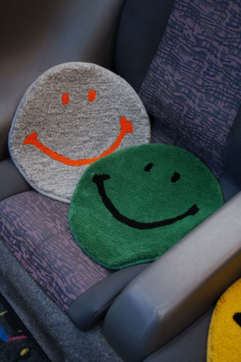 The MIYOSHI RUG Harvey Ball x MIYOSHI RUG Original Smiley Face Chair Rug features two handmade, Japan-inspired rugs: one gray with an orange smile and one green with a black smile, designed to resemble patterned bus or train seats.