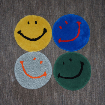 Four MIYOSHI RUG Harvey Ball x MIYOSHI RUG Original Smiley Face Chair Rugs—in yellow, blue, gray, and green—are arranged in a square on a brown striped carpet. Handmade in Japan, each features unique Harvey Ball-inspired facial details.