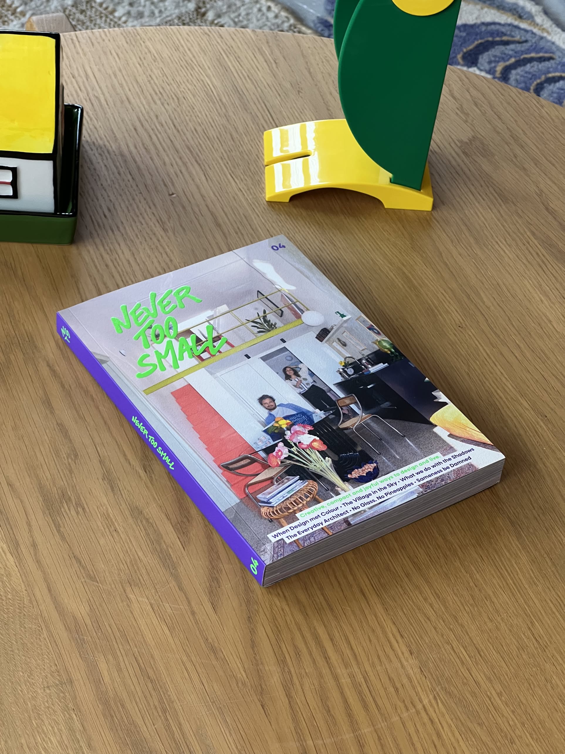 Never Too Small Magazine Issue #4 by Never Too Small, featuring a colorful modern cover, is displayed on a wooden table with decorative objects nearby.