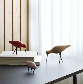 The Normann Copenhagen Shorebird Small Oak figurines by Normann Copenhagen in red, brown, and pink are displayed on a black table with books, bringing a touch of nature-inspired decor near a window with sheer curtains and wood panel walls.