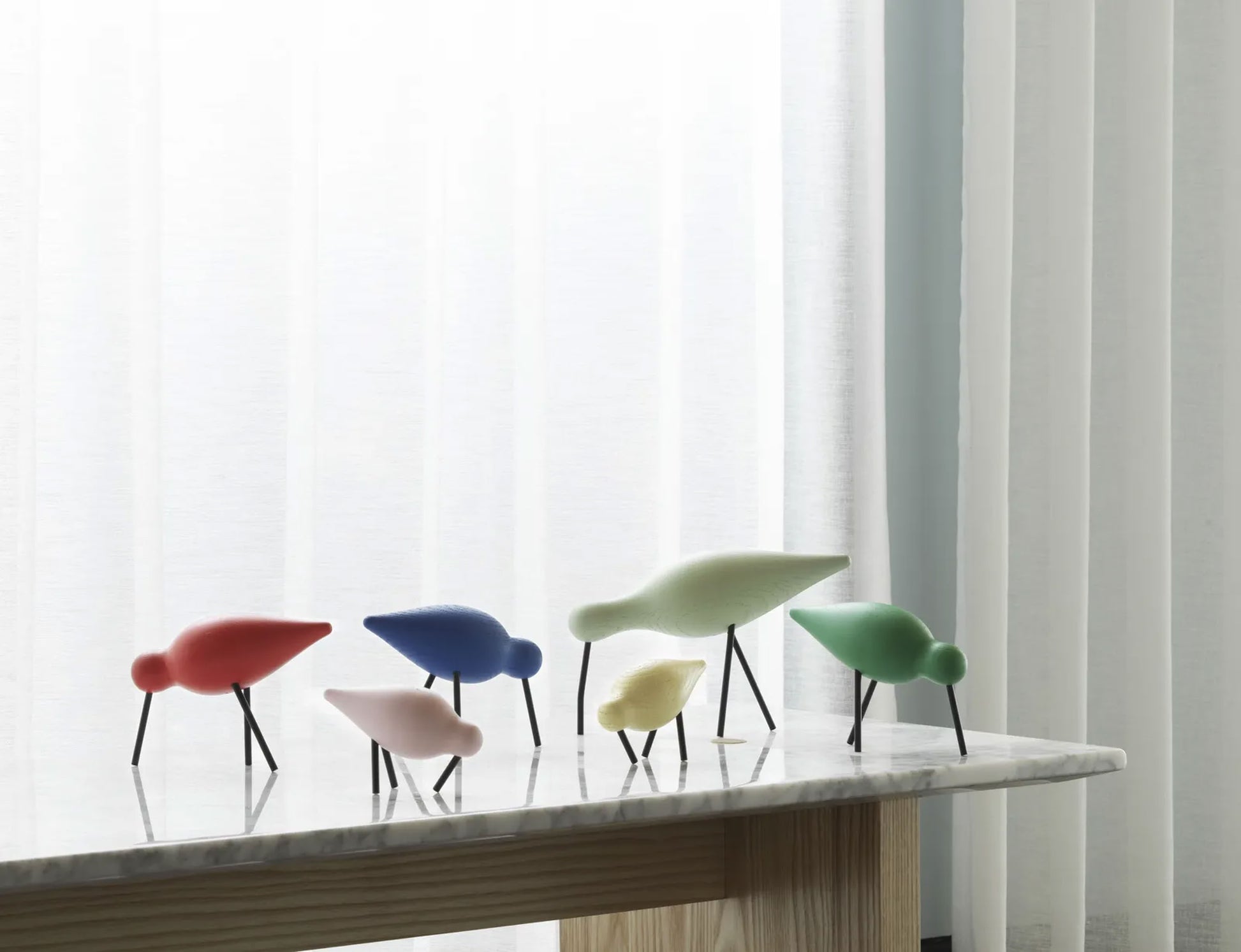 Five colorful Normann Copenhagen Shorebird Small Oak sculptures stand on a marble-topped table before white curtains. Designed by Normann Copenhagen, these oak birds add playful, minimalist charm in red, blue, pink, yellow, and green.