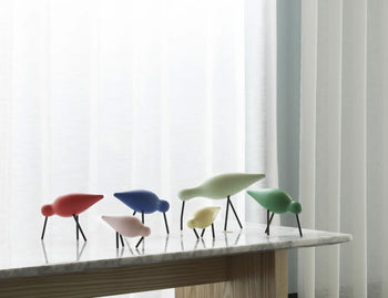 Five colorful Normann Copenhagen Shorebird Small Oak sculptures stand on a marble-topped table before white curtains. Designed by Normann Copenhagen, these oak birds add playful, minimalist charm in red, blue, pink, yellow, and green.